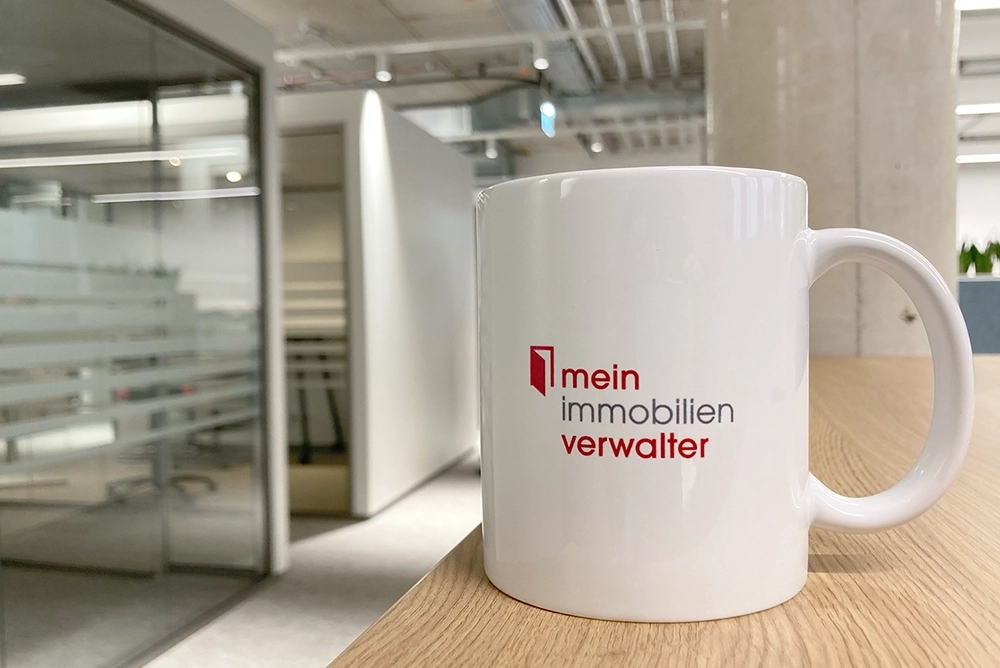 Eine Tasse mit dem Logo des Immobilienverwalters mit Büro im Hintergrund.